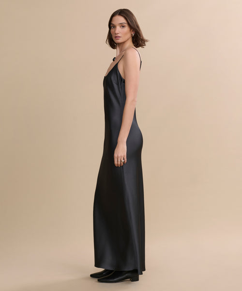 A woman stands sideways in the Cleo Slip Dress—long, black, with thin straps—paired with black boots against a beige background.
