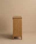 The Coast Hutch is a wooden cabinet with vertical slats, shown from the side against a beige background. Made from solid white oak with a light natural finish, it stands on tapered legs and provides stylish storage for any room.