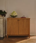 The Coast Hutch, crafted from solid white oak, stands against a beige wall with a glass vase of white flowers and a red tray holding two teacups on top. Next to it is a white marble fireplace adorned with floral arrangements.