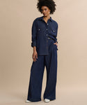 A woman with short curly hair and a confident expression stands against a beige background wearing the Western-inspired Colt Shirt in dark denim with matching wide-leg pants and white shoes, one hand in her pocket.