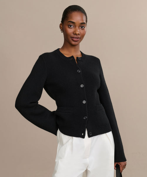A woman in the Cooper Cardigan, a structured black button-up, pairs it with white pants and stands against a beige background, looking at the camera with a slight smile.
