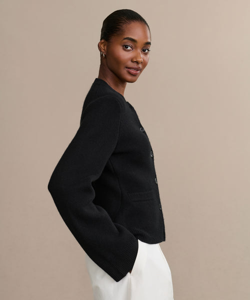A woman in profile stands against a beige background, wearing the fitted Cooper Cardigan. Her hair is pulled back and she smiles softly at the camera.