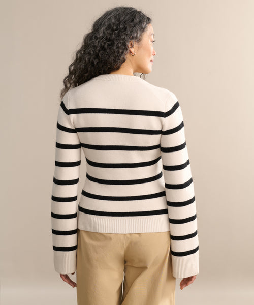 A woman with long, curly dark hair stands with her back to the camera, wearing the Cooper Cardigan in an ivory and black stripe pattern with beige pants, against a neutral background.