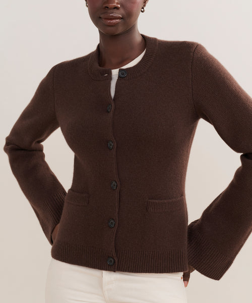 A person wears the Cooper Cardigan with a structured fit and two front pockets, styled over a white top and paired with white pants for a versatile look.