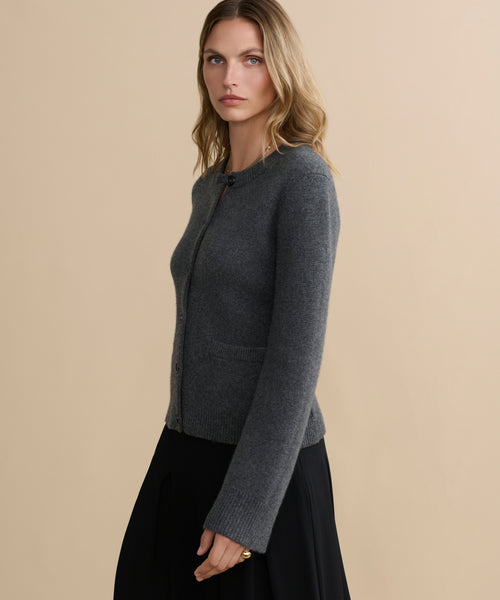 A woman with wavy blonde hair wears a fitted dark grey Cooper Cardigan and a black skirt, standing against a beige background and looking at the camera with a neutral expression.