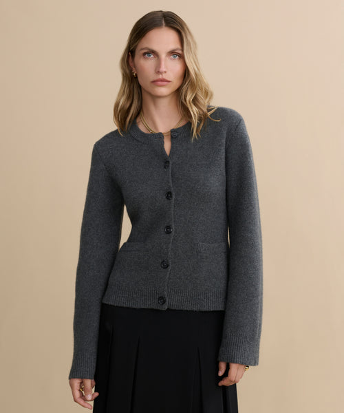 A woman with wavy blonde hair wears the fitted dark grey Cooper Cardigan and a black skirt, standing against a plain beige background and looking directly at the camera with a neutral expression.