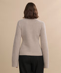 A person with shoulder-length brown hair, dressed in the Cooper Cardigan in light beige and black pants, stands with their back to the camera against a plain beige background.