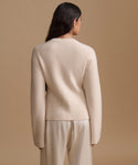 A woman shown from the back wearing a light cream knit sweater and matching trousers against a solid beige studio background.