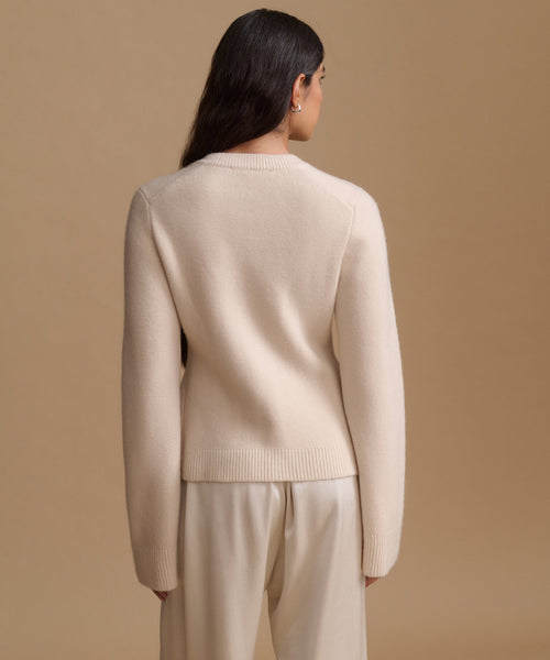 A woman shown from the back wearing a light cream knit sweater and matching trousers against a solid beige studio background.