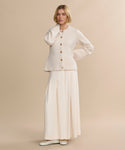 A woman with short blonde hair wears a Cooper Cardigan in Warm Ivory, styled with a long cream skirt and cream shoes, standing against a plain beige background.