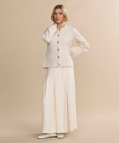 A woman with short blonde hair wears a Cooper Cardigan in Warm Ivory, styled with a long cream skirt and cream shoes, standing against a plain beige background.