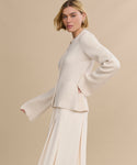 A woman with a blond bob wears the Cooper Cardigan in Warm Ivory, featuring wide sleeves and a matching long skirt, highlighting its structured silhouette as she poses against a plain beige background.