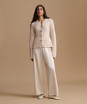 A woman stands against a warm beige studio background wearing a matching cream-colored sweater with long sleeves and wide-leg trousers, paired with cream flats.