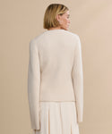 A woman with short blonde hair faces away from the camera, wearing the Cooper Cardigan in Warm Ivory and a matching pleated skirt, displaying a structured look against a soft beige background.