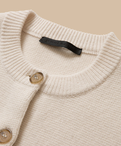 Close-up of the Cooper Cardigan, showcasing its cream knit in Warm Ivory with a ribbed collar, two beige buttons, structured shape, and a black fabric label at the neckline.
