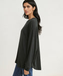 A woman with long dark hair stands sideways against a plain light background, wearing the Boyfriend Long-Sleeve Tee in black and blue jeans, looking at the camera.