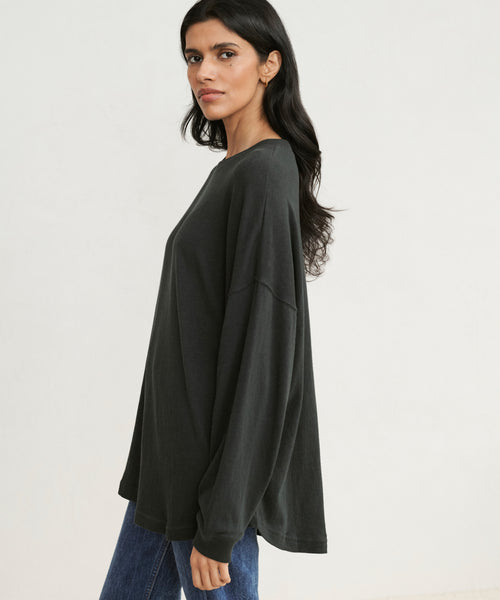 A woman with long dark hair stands sideways against a plain light background, wearing the Boyfriend Long-Sleeve Tee in black and blue jeans, looking at the camera.
