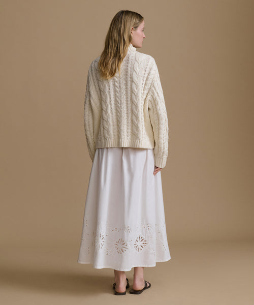 Woman wearing a cream-colored cable knit sweater over a white skirt on a beige background