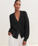 A woman stands against a plain light background, hand in pocket and relaxed, confidently wearing the Cotton Cocoon Cardigan—an organic cotton, loose-fitting black layer—paired with white pants.