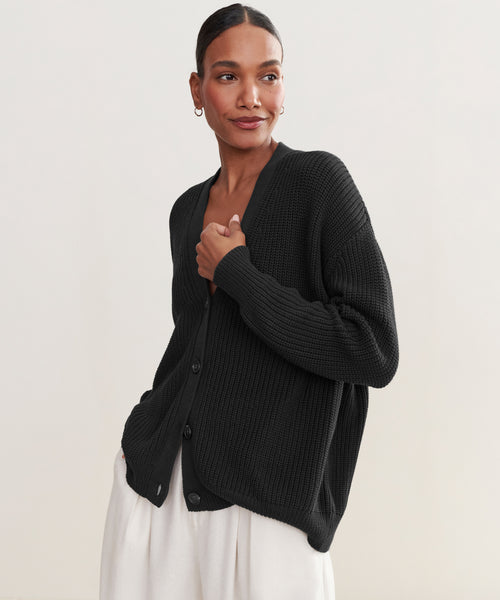 A woman models the Cotton Cocoon Cardigan in black with light pants, standing against a plain light background and looking to the side while holding the cardigan.