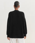 A person with hair in a low bun is seen from behind, wearing the Cotton Cocoon Cardigan in black with loose white pants against a light background.