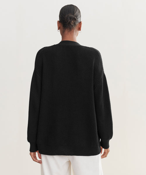 A person with hair in a low bun is seen from behind, wearing the Cotton Cocoon Cardigan in black with loose white pants against a light background.