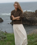 Woman wearing a brown sweater and white skirt standing on a cliff overlooking the ocean.