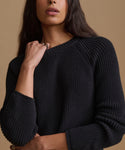 A woman modeling a black ribbed knit sweater and cream wide-leg trousers against a solid beige studio background.