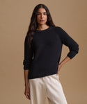 A woman modeling a black ribbed knit crewneck sweater with raglan sleeves, paired with cream-colored trousers, against a solid beige studio background.