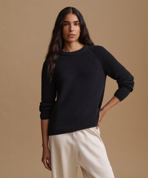 A woman modeling a black ribbed knit crewneck sweater with raglan sleeves, paired with cream-colored trousers, against a solid beige studio background.