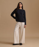 A woman modeling a black ribbed knit sweater and cream wide-leg trousers against a solid beige studio background.