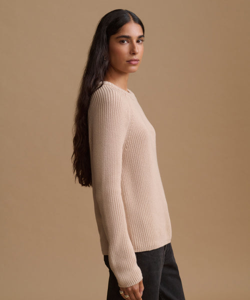 A woman shown in profile wearing a light beige ribbed knit sweater and dark pants against a solid beige studio background.