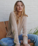 A woman sitting on a wooden bench wearing a beige ribbed knit sweater and blue jeans, set against a white brick wall with some green plants.
