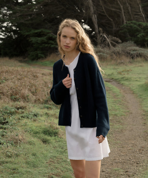 Woman in a navy cotton cardigan and white dress standing on a path in a natural setting