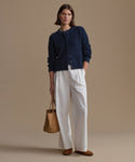 Woman wearing a navy cardigan, white pants, and brown shoes, holding a woven bag against a beige background.