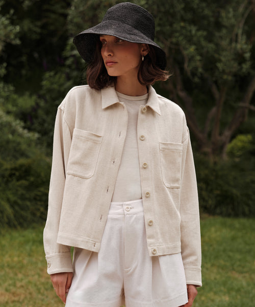 A woman stands outdoors among greenery, wearing the Georgia Jacket—a cream cotton linen piece—paired with a black wide-brim hat, white t-shirt, and white shorts.