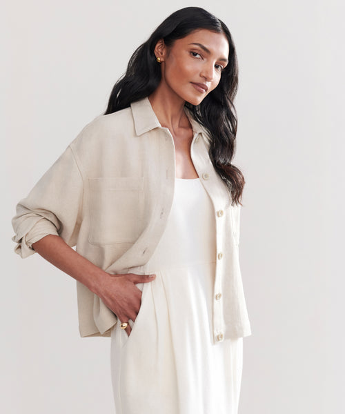 A woman with long, wavy dark hair stands against a light background wearing the Georgia Jacket over a white dress, one hand in her pocket and a relaxed, confident expression.