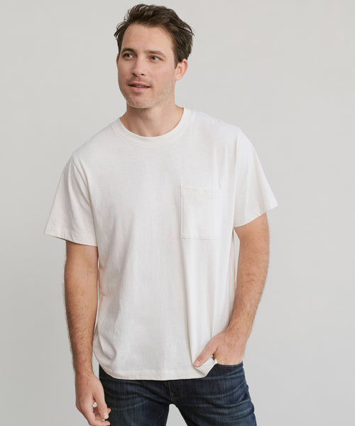A man stands against a plain background, wearing the Mens Vintage Pocket Tee in ivory with a chest pocket and dark blue jeans, showcasing its relaxed vintage fit as he looks slightly to the side with one hand in his pocket.