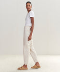 A woman stands sideways wearing a white t-shirt, light beige Saturday Sweatpant joggers, and tan slide sandals against a plain, light-colored background.