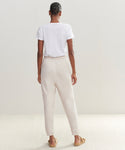 A person stands indoors facing away, wearing a white t-shirt, ultra-soft light beige Saturday Sweatpant made from French terry, and tan slippers against a plain neutral background.