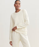 Someone models the cream Saturday Sweatshirt, made of soft French terry, paired with matching sweatpants. They stand against a plain, light background and look confidently at the camera, one hand on their hip.