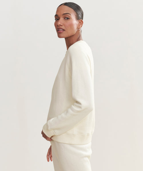 A person with slicked-back hair models the light cream Saturday Sweatshirt and matching soft French terry pants, standing sideways and facing the camera against a plain background.