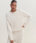 A woman stands against a light background, smiling softly. She wears the Saturday Sweatshirt in light beige French terry with matching sweatpants, her right hand on her hip and left arm relaxed by her side.