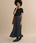 Wearing a Cotton Slim Tee with a fitted silhouette, a long pleated black skirt, and black boots, a woman stands against a beige background holding a camel coat over her shoulder.