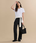 A woman stands against a beige background wearing the Cotton Slim Tee in white, black pants, and shoes, with a cream sweater tied at her waist. She holds a black handbag and touches her head with one hand.