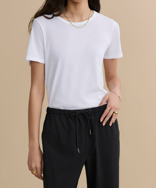 A woman wearing the Cotton Slim Tee and black drawstring pants stands against a beige background. She has long dark hair and accessorizes with gold jewelry, including a necklace and rings.