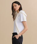 A woman with long, wavy dark hair stands sideways, wearing the Cotton Slim Tee in white, paired with black pants and a cream sweater over her shoulders. She has a neutral expression and stands against a minimal background.
