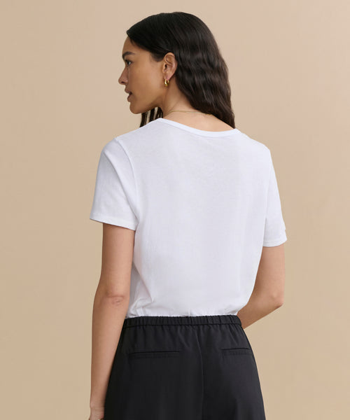 A woman with long dark hair stands with her back to the camera against a beige background, wearing the Cotton Slim Tee and black pants.