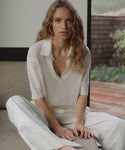 Woman in a ivory courtney top sitting on a couch in a room with a window view.