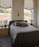 A cozy bedroom features a neatly made bed with a Cove Bed Slipcover, two beige pillows, matching bedside tables and lamps, large windows with natural light, artwork between the windows, and trees visible outside.